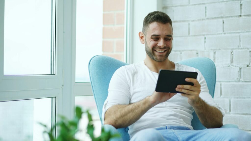 Happy man sitting in blue chair indoors, using a tablet, bright natural lighting.